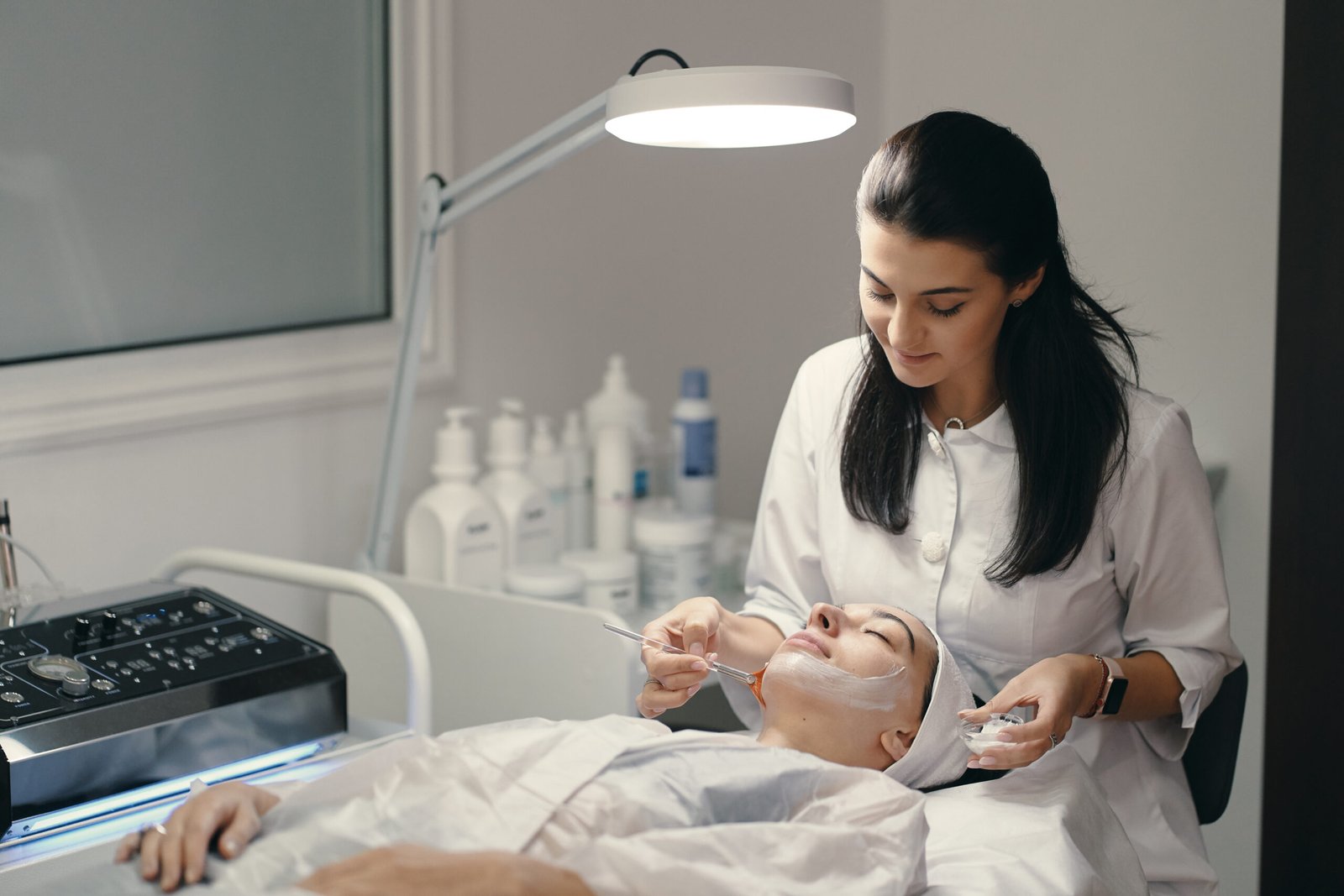 People, beauty, spa, cosmetology and skin care concept - Cosmetologist applying cream on a woman beautiful face with a cosmetologist brush. Lamp light focused on face.